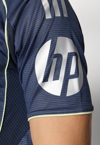 Maglia sportiva blu scuro con tessuto texturizzato, dotata di rifiniture verde lime e grande logo HP bianco sulla manica. Tre strisce bianche sopra.