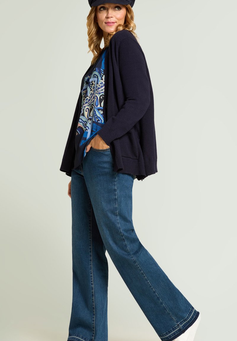 Marineblauer Cardigan über einem blau gemusterten Hemd, kombiniert mit ausgestellten Jeans und weißen Sneakers, dazu eine dunkle Mütze. Vorstellung eines lässigen Outfits.