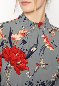 Bluzka w kwiatowy wzór na szarym tle, z czerwonymi i kremowymi kwiatami oraz marszczonym dekoltem o teksturowanym wykończeniu.