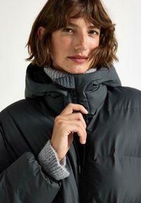 Nainen, joka pitää päällään tummaa untuvatakkia ja harmaata neulepaitaa, vetää takin vetoketjua ylös leukansa lähelle, vaalea tausta.