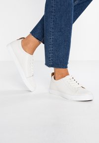 Valkoiset nahkakengät, joissa on sileä pinta, nauhat, erottuva ruskea kantapääkoristus ja valkoinen kumipohja.