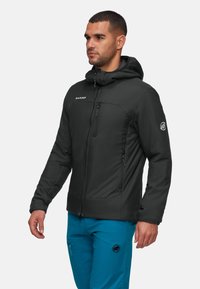 Uomo in giacca con cappuccio nera e pantaloni blu, in piedi e che guarda di lato su uno sfondo semplice.