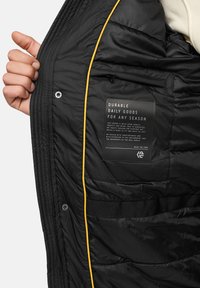 Zwart gewatteerd jasje aan de binnenkant met gele piping, voorzien van een stoffen label en een ritszak. Het materiaal is glad en licht glanzend.