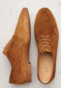 Chaussures en daim marron à lacets avec un bout pointu, perforations décoratives sur les côtés et une semelle en caoutchouc marron lisse.