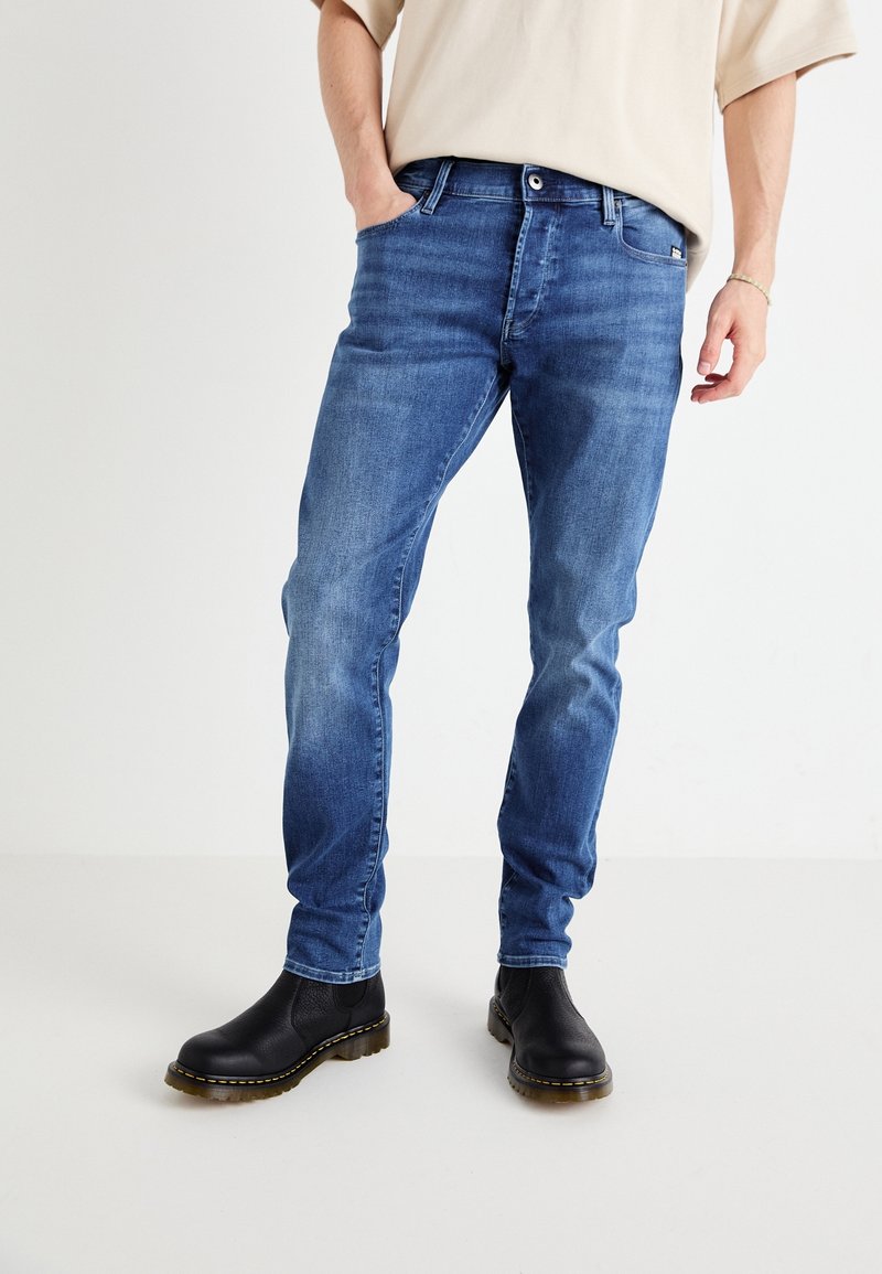 Homme portant un jean slim bleu moyen et des bottines en cuir noir, debout avec une main dans la poche devant un fond uni.