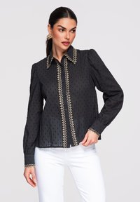 Donna che indossa una camicetta nera a maniche lunghe con rifiniture dorate e pantaloni bianchi, in piedi davanti a uno sfondo neutro, che guarda di lato.