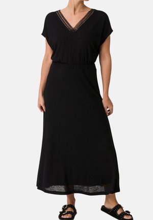 Frau trägt ein schwarzes Maxikleid mit V-Ausschnitt, Spitzenbesatz und elastischer Taille, kombiniert mit schwarzen Slip-On-Sandalen mit Nieten.