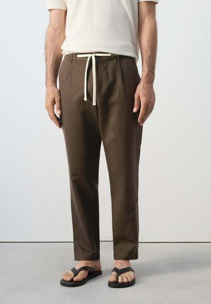 Mann trägt braune Kordhose und schwarze Flip-Flops und steht vor einem schlichten hellen Hintergrund.