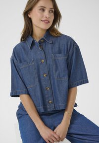 Afskåret denimskjorte i mellemblå, med to frontlommer, korte ærmer og brune knapper. Glat tekstur med synlig søm.