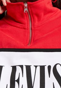 Rode fleece zip-up top met een hoge kraag. Bevat een witte paneel eronder met gedurfde zwarte tekst. Zichtbare zilveren rits en gestructureerde stof.