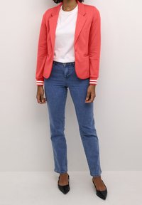 Koralowa marynarka z klapami z wcięciem i paskami na mankietach, zestawiona z białym t-shirtem i wysokim stanem niebieskich dżinsów, uzupełniona czarnymi spiczastymi butami.