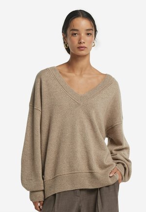 Beżowy, oversize sweter z dekoltem w serek, obniżonymi ramionami, ściągaczami przy mankietach i hemie. Miękka tekstura z subtelnym wzorem, stylizowany z luźnymi spodniami.