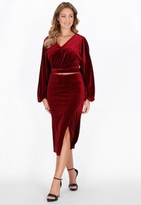 Piros bársony kétdarabos szett, amely egy hosszú ujjú, rövid felsőből és egy magas hasítékú midi szoknyából áll. A megjelenést fekete pántos magassarkú cipő teszi tökéletessé.