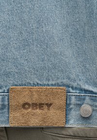 Τζιν ύφασμα σε ανοιχτό γαλάζιο με υφασμένη καφέ δερμάτινη ετικέτα με την ένδειξη "OBEY" και μεταλλικό κουμπί κοντά στην άκρη.