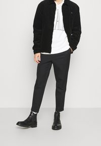 Giacca di velluto a coste nera, t-shirt grafica bianca, pantaloni neri e stivali alla caviglia neri. La giacca presenta un colletto e polsini elasticizzati.