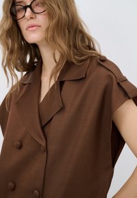 Blazer marrone senza maniche con colletto a risvolto, caratterizzato da bottoni grandi e un design strutturato. Il tessuto appare liscio e leggero.