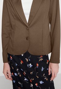 Blazer ajustado marrón con dos botones sobre una blusa blanca, combinado con una falda negra con un patrón floral multicolor.