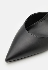 Svart läder sko med spetsig tå, som har en slät yta och en design med öppen sida, med sömdekorationer längs kanterna.
