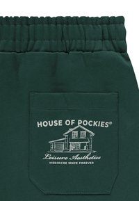 Zielone spodnie dresowe z marszczonym paskiem w talii, dużą tylną kieszenią z białym nadrukiem przedstawiającym grafikę domu oraz napisem "HOUSE OF POCKIES" powyżej.
