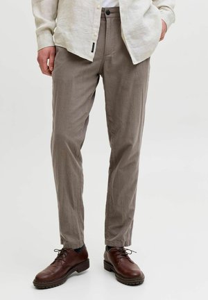 Mężczyzna ubrany w proste spodnie w kolorze taupe, brązowe skórzane buty na sznurowadła oraz jasnobeżową koszulę zapinaną na guziki, z jedną ręką w kieszeni.