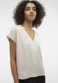 Blouse beige clair en V à manches courtes, fabriquée en tissu lisse et texturé ; coupe décontractée, avec un éclat subtile et une texture douce.