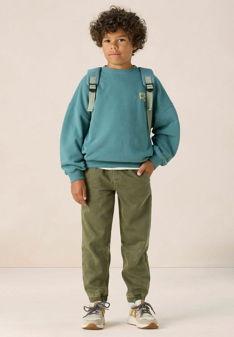 Sweatshirt surdimensionné turquoise avec des poignets côtelés, pantalon tapered vert olive, et basket beiges avec des accents gris et bleu. Bretelles réglables.