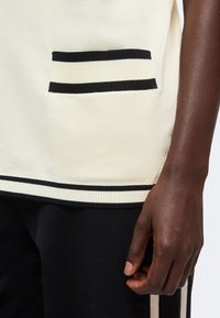 Main à la peau foncée reposant sur le côté portant un pantalon noir avec une bande blanche, à côté d’un haut en maille blanc avec une poche et un ourlet rayés noirs.