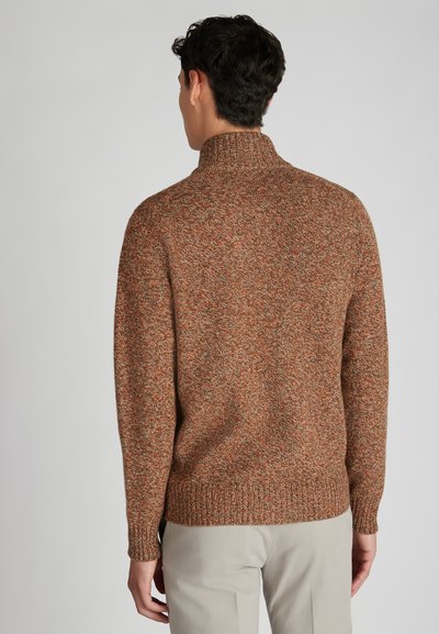 Brązowy sweter dziergany z wysokim kołnierzem, z teksturowanym wzorem. Żebrowane mankiety i dół dodają szczegółów. Model ma na sobie jasne spodnie.