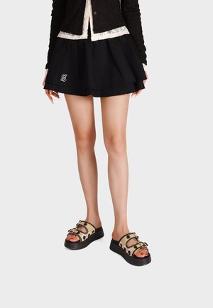 Donna che indossa una gonna nera a strati e sandali beige con plateau e motivo floreale nero, in piedi su uno sfondo bianco semplice.