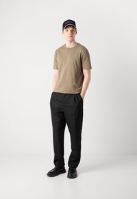 Nuori mies, jolla on khaki T-paita, mustat housut, mustat nahkakengät ja musta BOSS-lippis, seisoo kädet taskuissa valkoista taustaa vasten.