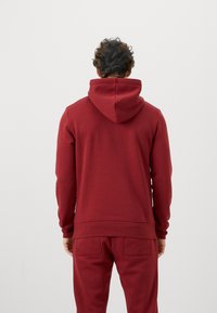 Muž s kučeravými vlasmi, oblečený v jednoduchom červenom mikinovom saku a zodpovedajúcich červených teplákoch, stojí chrbtom k fotografii na jednoduchom pozadí.