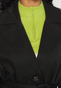 Trench-coat noir cintré à la taille, avec un grand col et un bouton noir proéminent. En dessous, un haut vert vif côtelé à col haut.