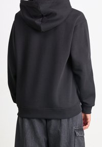 Felpa nera con cappuccio, tessuto liscio, vestibilità comoda e polsini e orlo a costine. Dotata di tasca a marsupio e cappuccio con coulisse regolabile.