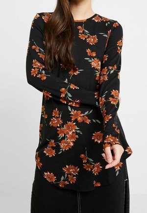 Vrouw in een zwarte langmouwen top met oranje bloemenprint, gecombineerd met een zwarte rok met witte stikdetails.