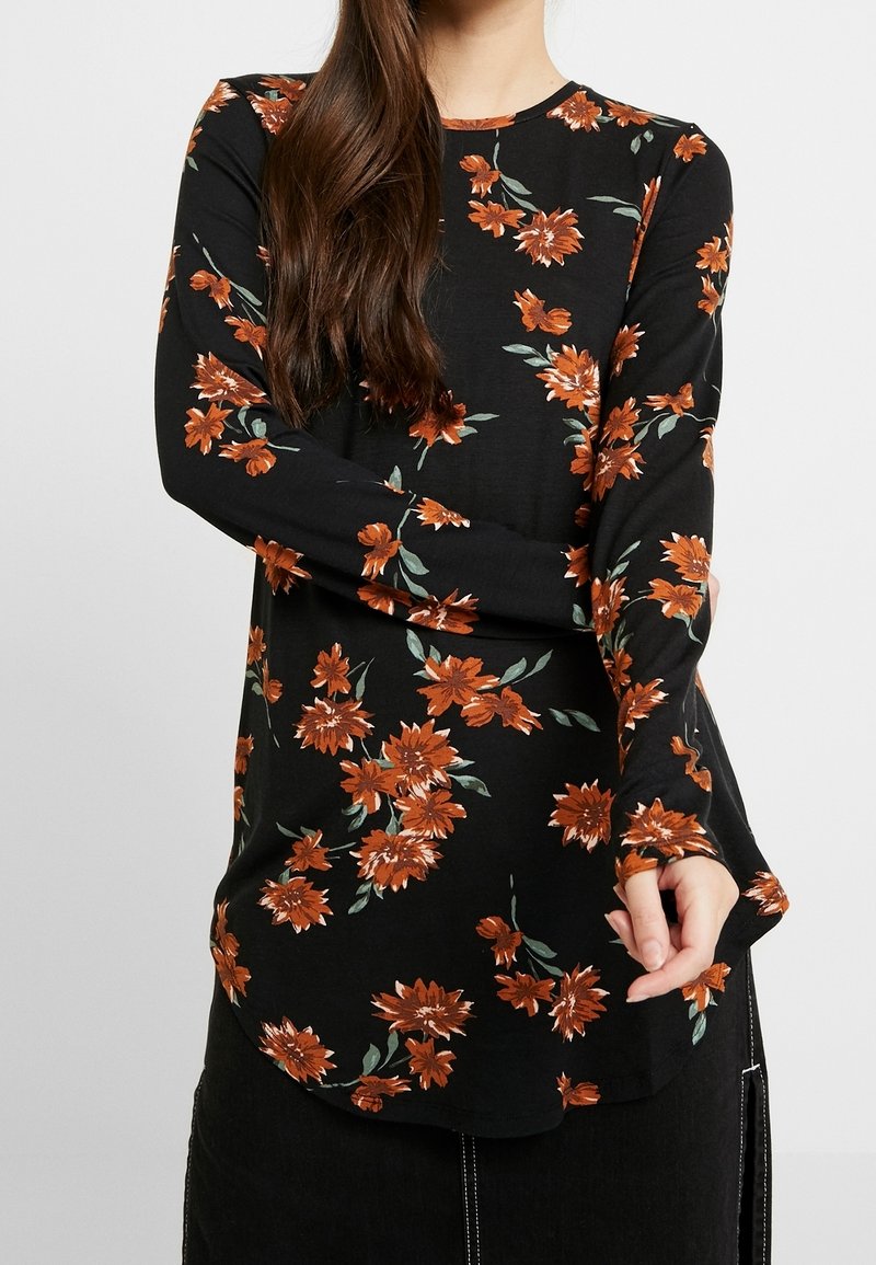 Mujer vistiendo una blusa de manga larga negra con estampado floral naranja, combinada con una falda negra con detalles de costura blanca.