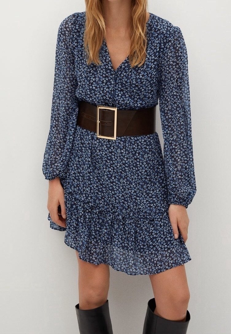 Une femme porte une robe longue à manches longues bleue à fleurs avec une large ceinture noire et des bottes noires montantes sur un fond clair uni.