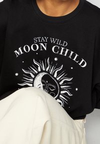 Sudadera negra con un gráfico del sol y la luna y el texto "STAY WILD MOON CHILD" en blanco. Tela suave y corte relajado.