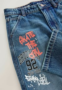 Mėlynos skalbimo džinsai su raudonu graffiti tekstu "SKATER" ir baltomis detalėmis. Įtraukti spausdinti užrašai "BROOKLYN 92" ir "SUPER COOL."