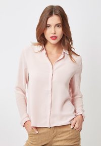 Blusa de manga larga en un suave tono rosa claro, con cuello, cierre de botones en la parte delantera y textura lisa. La tela parece ligera y suave, adecuada para diversas ocasiones.