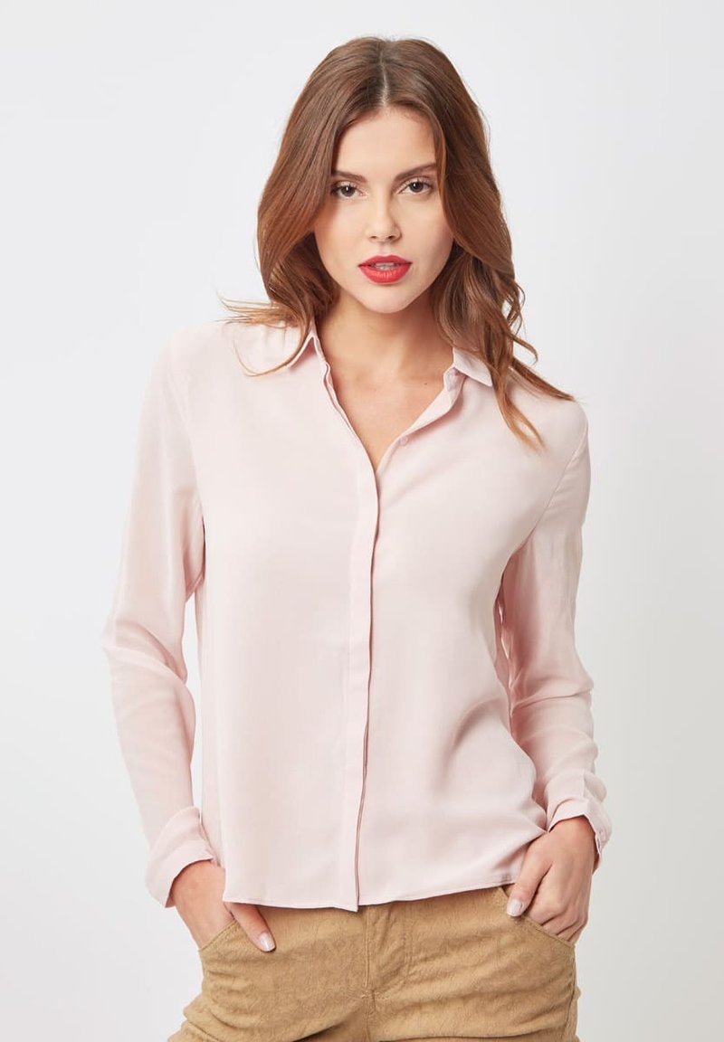 Blusa de manga larga en un suave tono rosa claro, con cuello, cierre de botones en la parte delantera y textura lisa. La tela parece ligera y suave, adecuada para diversas ocasiones.