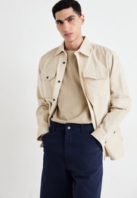 Beige cotton shirt jacket with two chest pockets, worn over a tan T-shirt and navy trousers. Textured fabric and subtle details.
