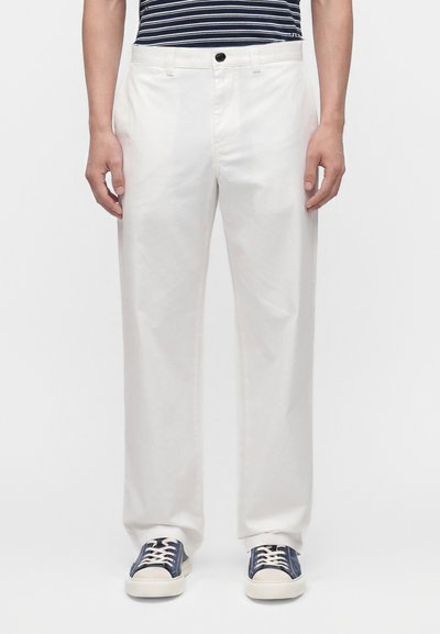 Homme portant un pantalon blanc droit, des baskets bleu marine avec des lacets blancs et une chemise rayée bleu marine, debout devant un fond uni.