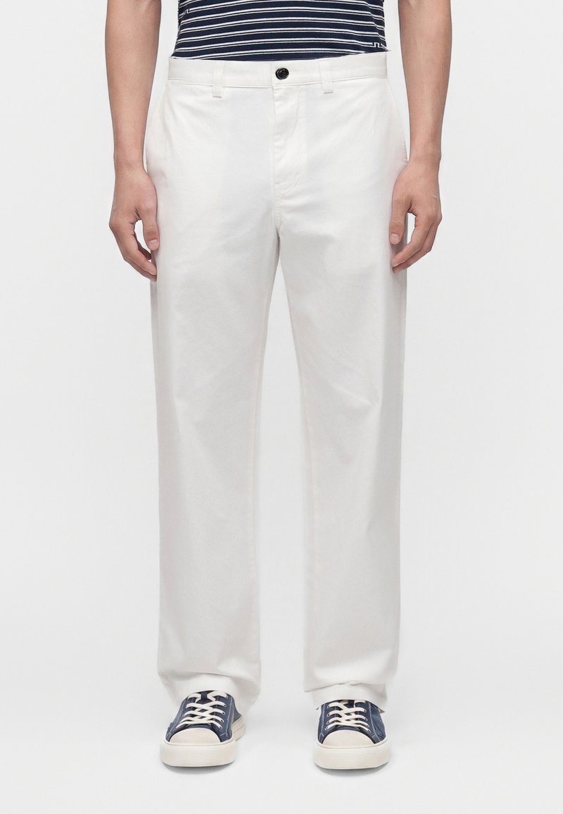 Homme portant un pantalon blanc droit, des baskets bleu marine avec des lacets blancs et une chemise rayée bleu marine, debout devant un fond uni.