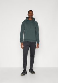 Tmavě zelená mikina s kapucí a šňůrkou, kombinovaná s černými slim-fit kalhotami. Oblečení má decentní logo na hrudi a u kotníků.