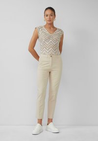 Pantalons beiges ajustés avec une fermeture à bouton associés à un haut à motifs à manches courtes et décolleté en V dans des teintes dorées et blanches, portés avec des baskets blanches.