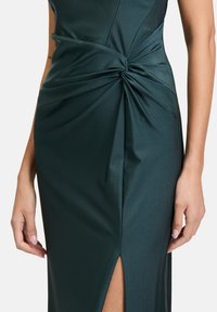Dunkelgrünes Satin-Kleid mit einem verdrehten Knoten-Detail an der Taille und einem Seitenschlitz, der die glatte Textur und das figurschmeichelnde Design betont.