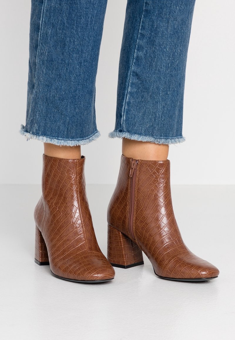 Bottines en cuir à texture crocodile marron avec des talons carrés portées avec un jean bleu à ourlet effiloché sur un sol clair.