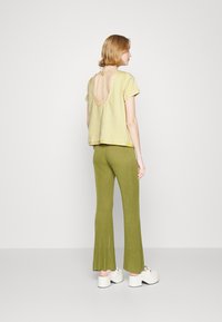 Top amarelo de manga curta com costas abertas e detalhe de laço, combinando com calças verde caneladas e flare. Modelo está a usar sapatos brancos de plataforma.
