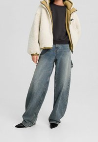 Person iført brede blå jeans, sorte spidse sko, sort top og cremfleecejakke med tan-linning, der står foran en ensfarvet baggrund.