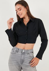 Black cropped button-up shirt with a pointed collar and long sleeves. Fabric is smooth with a structured fit. Paired with grey high-waisted jeans.